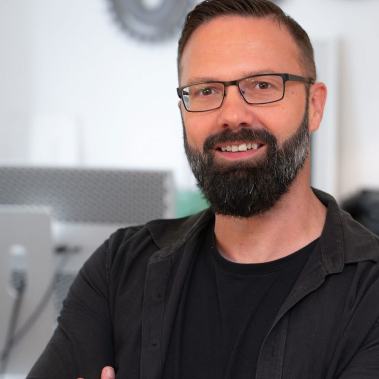 Jochen Riecke, Videograf und Spezialist für Filmproduktionen aus Rahden, steht locker im Büro von Seite an Seite. Er trägt ein schwarzes Shirt und eine schwarze Hemdjacke. Im Hintergrund sind verschwommen große Zahnräder als Wanddeko und ein Bildschirm erkennbar.