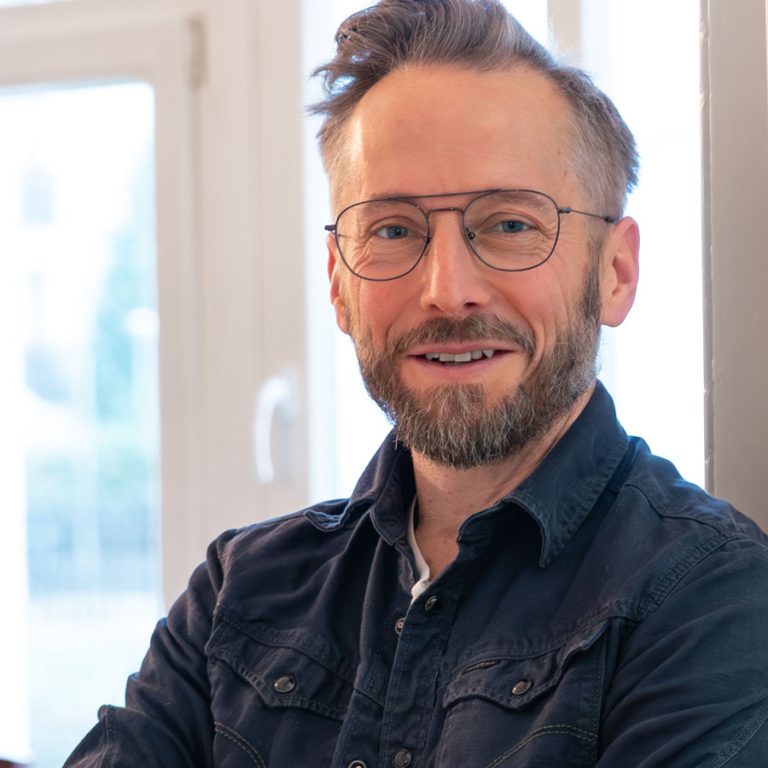 Oliver Windus, Fotograf und Gründer von Windus Fotodesign aus Löhne, steht mit leicht geneigtem Kopf und verschränkten Armen vor einer hellen Fensterfront. Er trägt eine dunkle Jeansjacke und eine randlose Brille, sein Blick ist offen und freundlich.