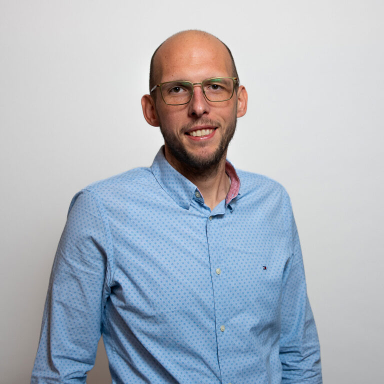 Porträt eines lächelnden Mannes mit Brille, der ein hellblaues Hemd trägt und vor neutralem grauem Hintergrund steht.