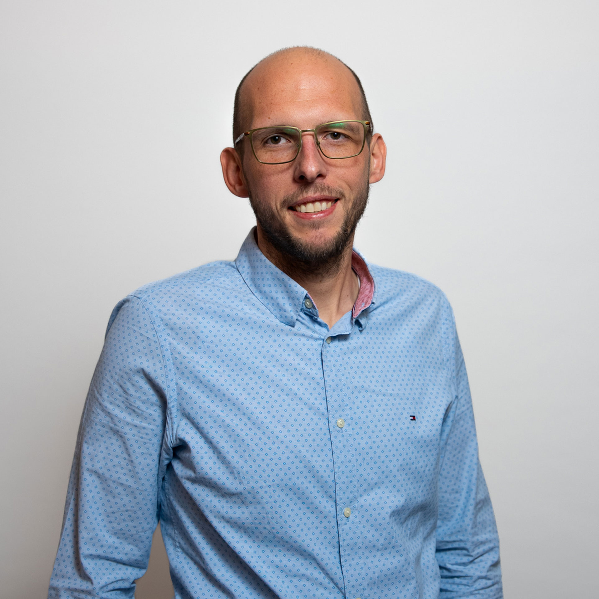 Porträt eines lächelnden Mannes mit Brille, der ein hellblaues Hemd trägt und vor neutralem grauem Hintergrund steht.