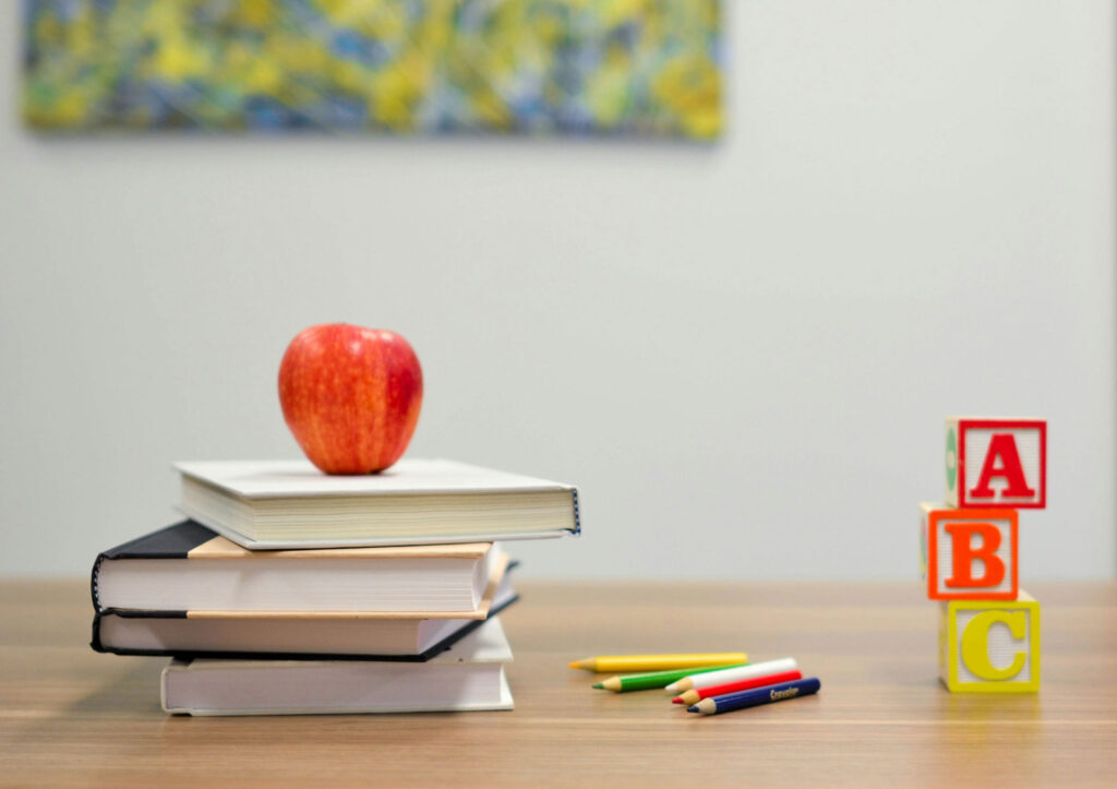 Ein roter Apfel liegt auf einem Stapel aus vier Büchern auf einem Holztisch. Daneben liegen bunte Stifte, rechts sind drei Bauklötze mit den Buchstaben A, B und C gestapelt. Im Hintergrund ist ein unscharfes, farbenfrohes Bild zu sehen.