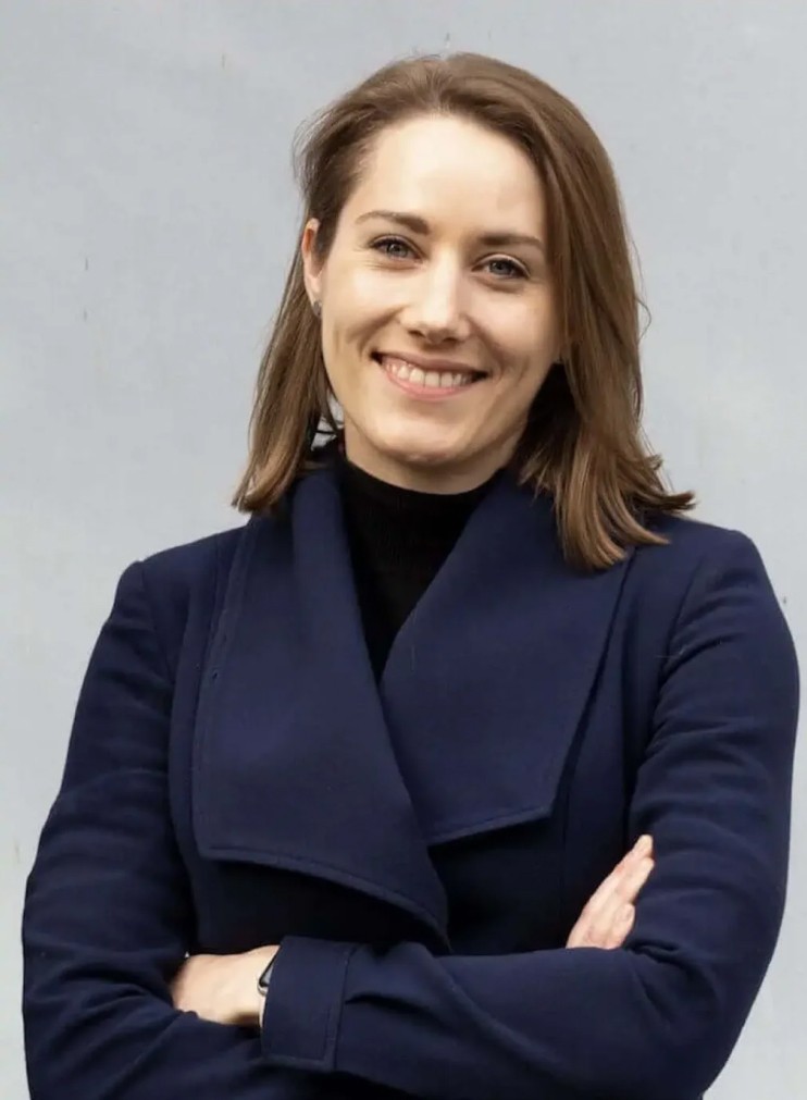 Portrait einer lächelnden Dame mit hellbraunen Haaren vor einer grauen Wand.