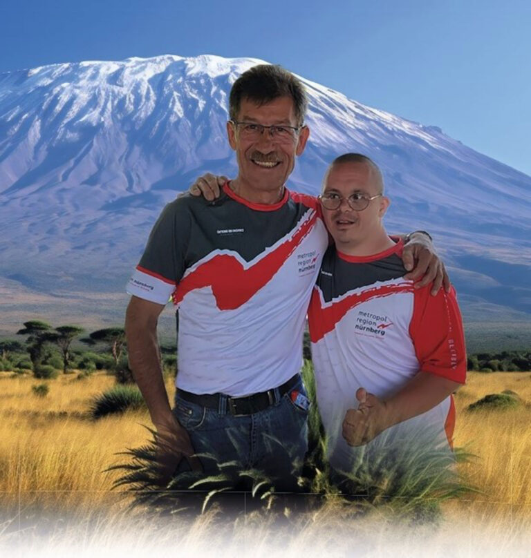 Extremsportler Bernhard Nuss steht mit seinem Freund Conny Arm in Arm vor dem Kilimandscharo. Beide tragen rot-weiße Sportshirts und lächeln in die Kamera. Im Hintergrund sind die schneebedeckten Hänge des Berges sowie eine weite Savannenlandschaft zu sehen.