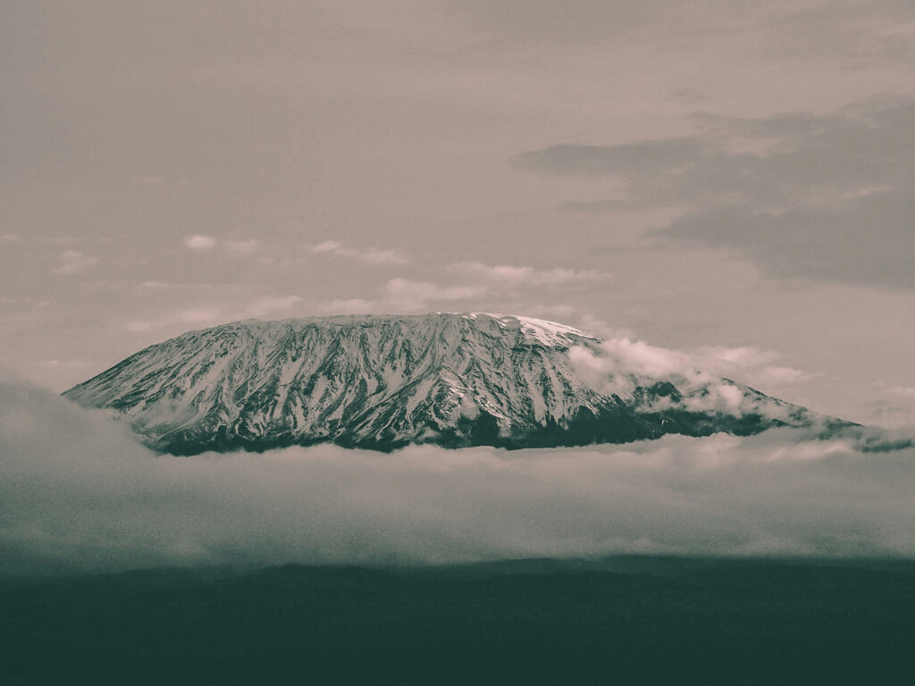 Der schneebedeckte Kilimandscharo ragt über einer dichten Wolkendecke empor. Sein breiter Gipfel ist von dunklen Felsstrukturen und hellen Schneefeldern geprägt, während tief hängende Wolken Teile des Bergmassivs verhüllen. Die Aufnahme wirkt ruhig, kühl und atmosphärisch.