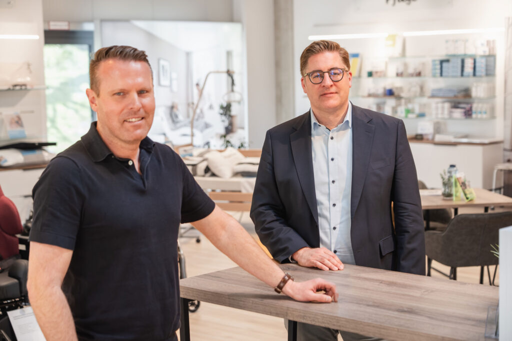 Elmar Bitter und Thorben Grünewälder stehen während eines Interviews für Seite an Seite in den hellen Geschäftsräumen von Orthopartner an einem Beratungstisch.