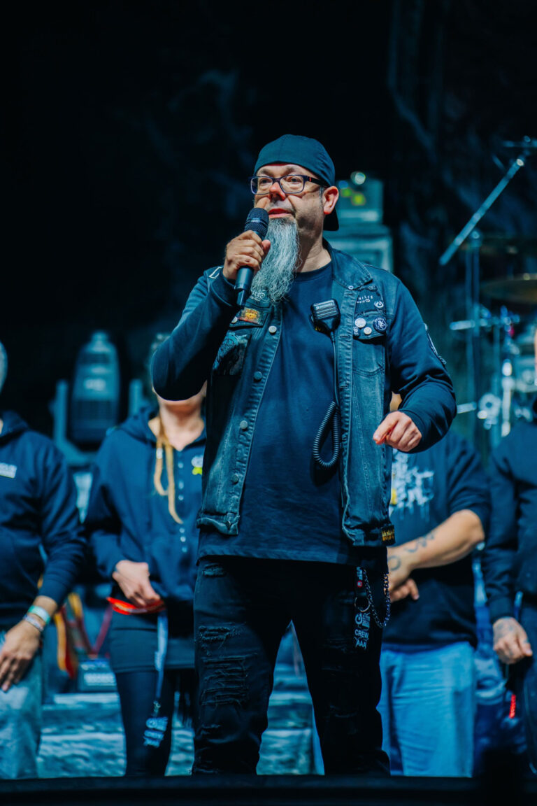 Marco Spiller, Vorstandsvorsitzender der Lebenshilfe Braunschweig. Marco steht auf der Konzertbühne des inklusiven Festivals Rock in Rautheim und hält ein Mikrofon in der Hand und spricht zum Publikum. Marco trägt eine Jeansweste und eine Mütze. Er wirkt freundlich und begeistert. Im Hintergrund stehen Mitglieder des Teams der Lebenshilfe Braunschweig, die das Festival organisert. Das Bild begleitet ein Interview mit Seite an Seite, dem Netzwerk für barrierefreies Marketing.