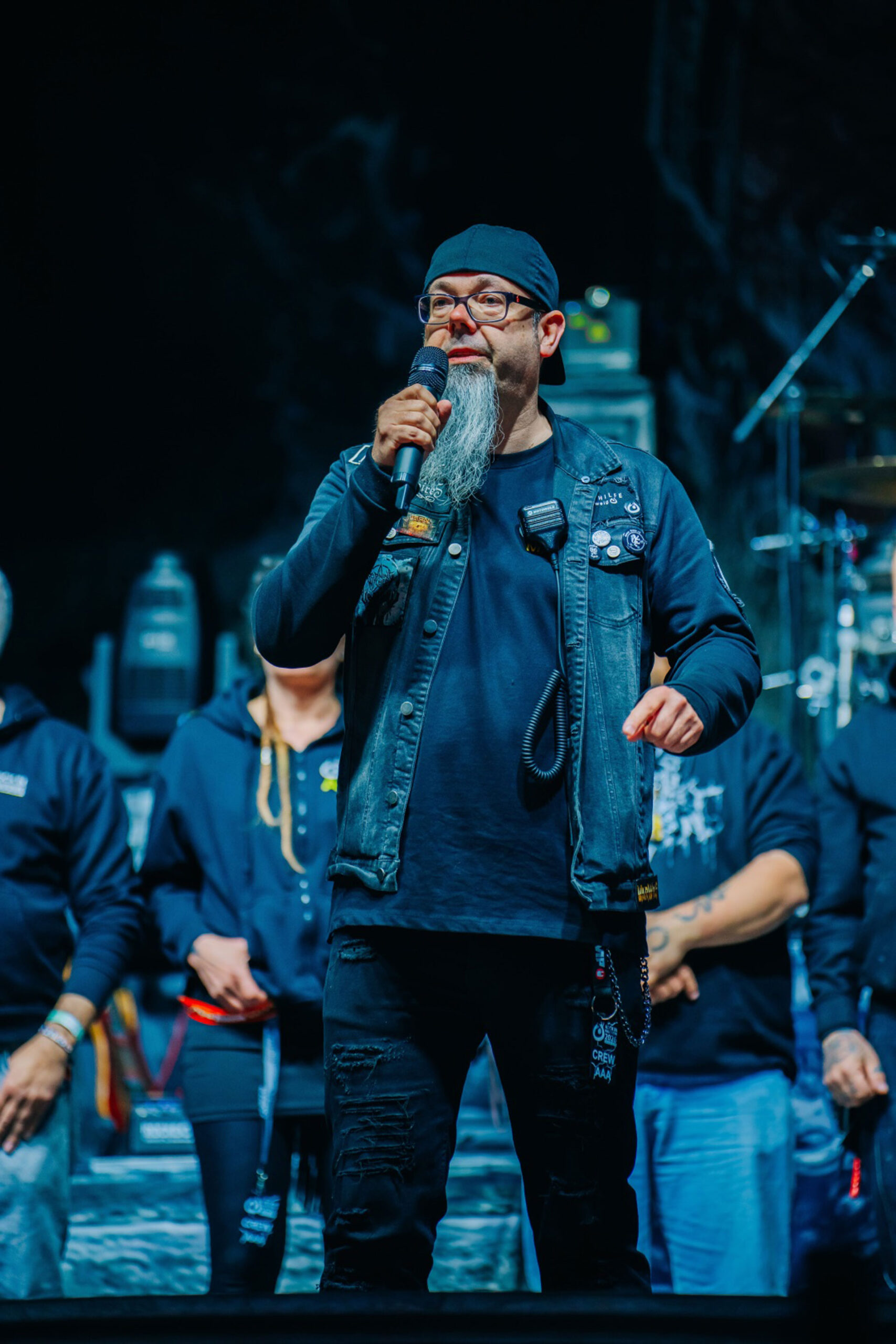 Marco Spiller, Vorstandsvorsitzender der Lebenshilfe Braunschweig. Marco steht auf der Konzertbühne des inklusiven Festivals Rock in Rautheim und hält ein Mikrofon in der Hand und spricht zum Publikum. Marco trägt eine Jeansweste und eine Mütze. Er wirkt freundlich und begeistert. Im Hintergrund stehen Mitglieder des Teams der Lebenshilfe Braunschweig, die das Festival organisert. Das Bild begleitet ein Interview mit Seite an Seite, dem Netzwerk für barrierefreies Marketing.