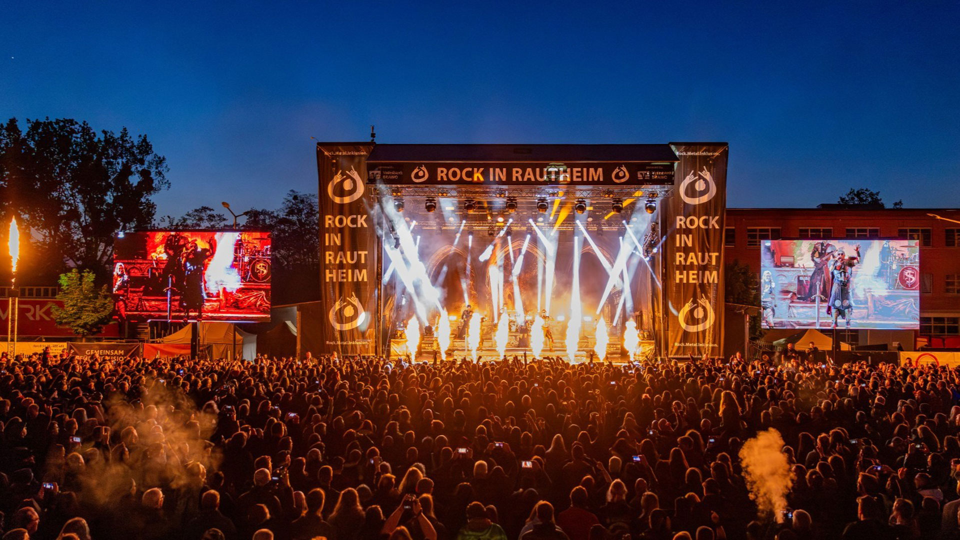 Lichtshow und Pyrotechnik auf der Bühne beim inklusiven Festival Rock in Rautheim vor großem Publikum. Auf der Bühne spielt die Rockband Feuerschwanz. Das Publikum steht vor der Bühne und feiert die Band. Es ist spät am Abend und die Pryotechnik der Band beleuchtet die Szenerie.